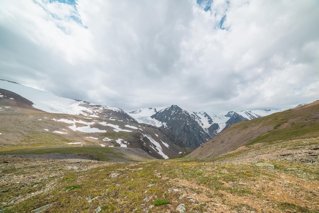 Vista panorámica escénica desde la colina cubierta de hierba verde iluminada por el sol hasta la alta cordillera nevada con cimas afiladas y glaciares bajo un cielo nublado gris Paisaje colorido con grandes montañas nevadas en un clima cambiante