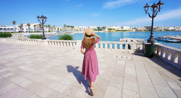 Vista panorámica de la hermosa joven con sombrero y vestido caminando por el paseo marítimo de Otranto, Puglia, Italia