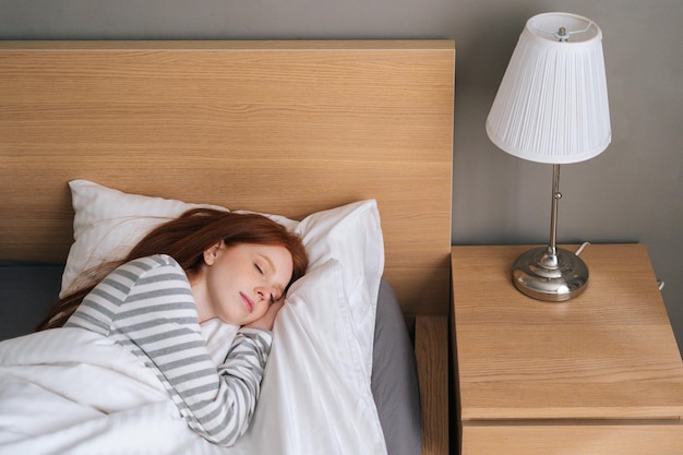 Vista panorámica de una joven pelirroja bonita durmiendo bien en la cama abrazando una manta blanca suave en casa. Encantadora dama descansando disfrutando de ropa de cama fresca y suave y colchón en el dormitorio.