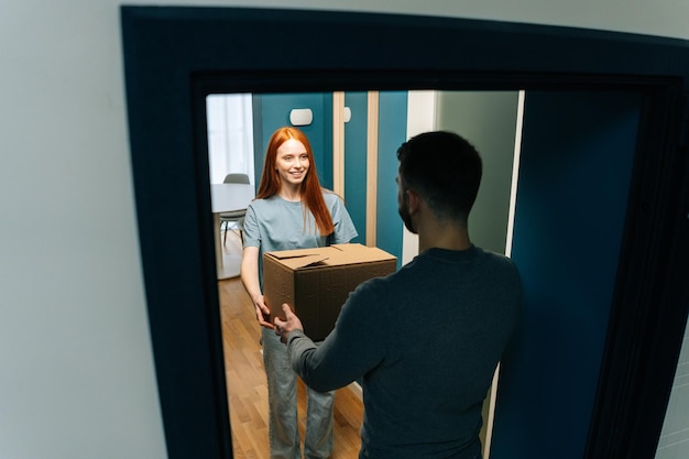 Vista panorámica de una joven que recibe una caja de cartón de un repartidor irreconocible en la puerta del apartamento