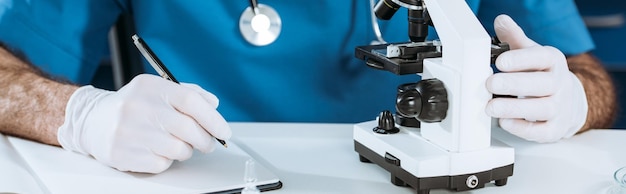 Vista parcial del biólogo en guantes de látex escribiendo en un cuaderno cerca de la toma panorámica del microscopio