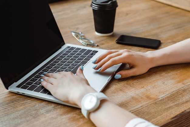 Vista parcial da mulher digitando no teclado do laptop perto de smartphone e café para ir à mesa de madeira no café