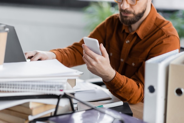 vista parcial de un hombre de negocios barbudo con gafas y camisa de moda sentado con un teléfono inteligente cerca