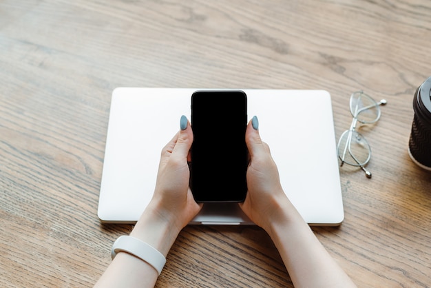 Foto vista parcial de la mujer que sostiene el teléfono inteligente con pantalla en blanco cerca de la computadora portátil en la cafetería