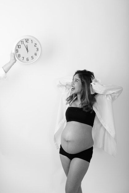 Vista de perfil de la joven mujer embarazada asiática feliz sonriendo mientras mira el reloj de pared y sosteniendo la cabeza