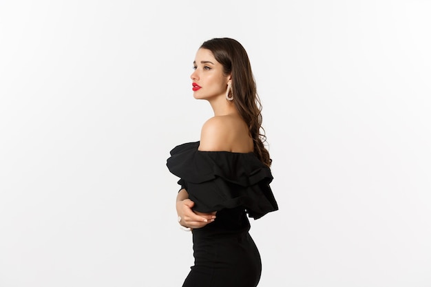 Vista de perfil de mujer joven elegante con labios rojos, maquillaje y vestido negro, mirando soñadora en la distancia, de pie sobre fondo blanco.