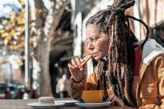 Vista de perfil de una mujer joven con rastas sentada afuera de la cafetería comiendo una galleta con trocitos de chocolate