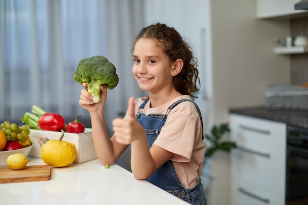 Vista de perfil de una niña con cabello rizado, vestida con jeans y camiseta, sosteniendo un brócoli, mostrando los pulgares, sentada a la mesa en la cocina.