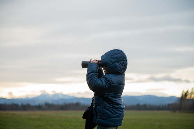 Vista de perfil de un niño pequeño emocionado de pie afuera en la hermosa naturaleza mirando a través de binoculares.