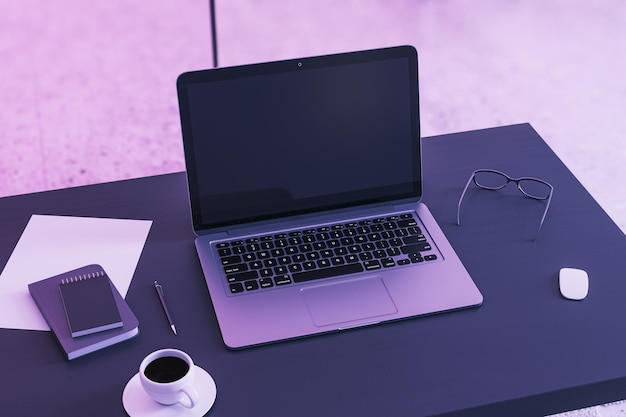 Vista en perspectiva superior en la pantalla del portátil moderno negro en blanco con lugar para el diseño web o página de destino en la mesa de trabajo oscura con taza de café y herramientas de oficina en la maqueta de renderizado 3D de fondo de pared púrpura