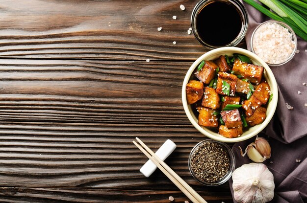 Vista plana en cubos de tofu salteados con cebollino en un plato de arcilla en la mesa de la cocina de madera