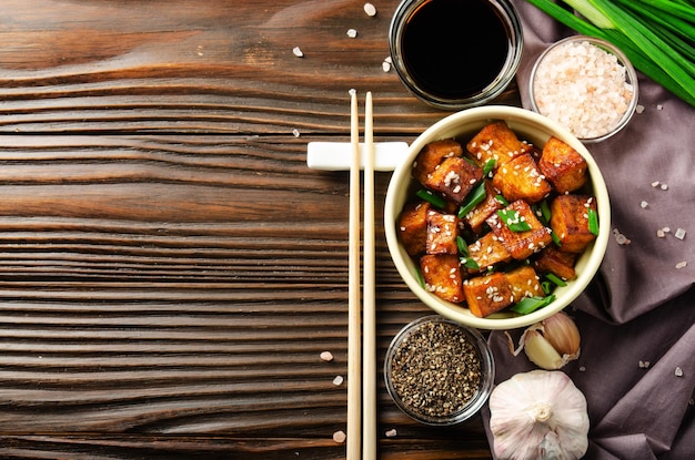 Vista plana en cubos de tofu salteados con cebollino en un plato de arcilla sobre una mesa de cocina de madera con una servilleta