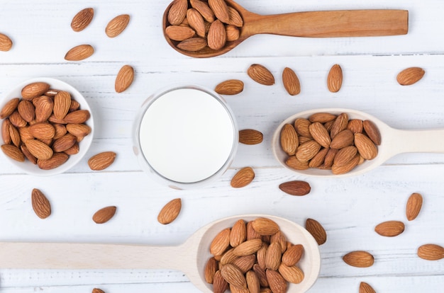 Vista plana de leche de almendras en vidrio con semillas y cucharas de madera en un plato de mesa de madera blanca