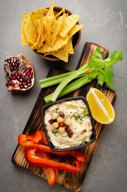 Vista plana del plato de hummus vegetal cubierto con garbanzos y aceite de oliva servido con chips de tortilla, apio y pimientos