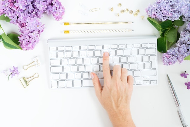Vista plana superior del espacio de trabajo de la oficina en casa escribiendo a mano en el teclado moderno con flores de color lila sobre fondo blanco