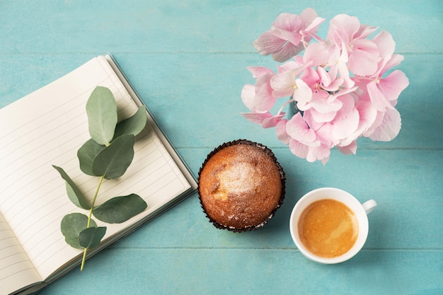 Vista plana, vista superior del escritorio femenino. Taza de café para el desayuno, magdalena, cuaderno vacío, rama con hojas de eucalipto y hortensia rosa. Concepto de desayuno de planificación empresarial.