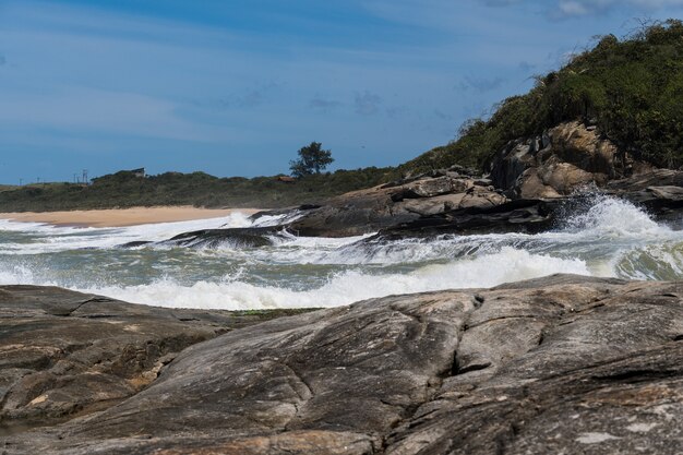 Vista de la playa de Areia Negras en Rio das Ostras en Rio de Janeiro con día soleado, cielo azul y algunas nubes. Mar fuerte y arena negra mezclada con amarillo y muchas rocas.