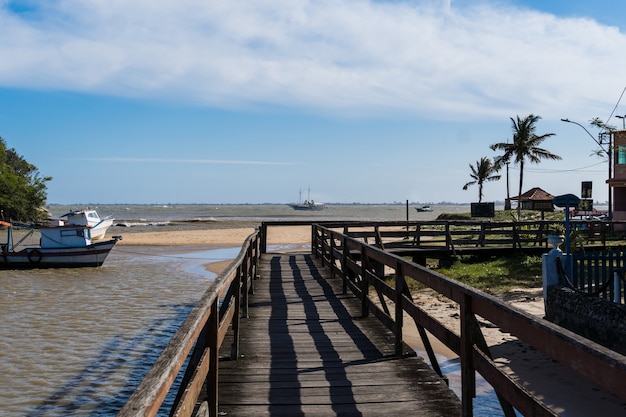 Vista de la playa de Rio das Ostras con el encuentro del río en Rio de Janeiro. Día soleado, cielo azul. Arena amarilla y algunas rocas. Puente de madera para cruzar. Barcos y pesca en el muelle.