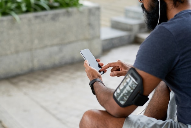 Vista por cima do ombro de um jovem indiano com barba sentado ao ar livre e usando o aplicativo de fitness no smartphone