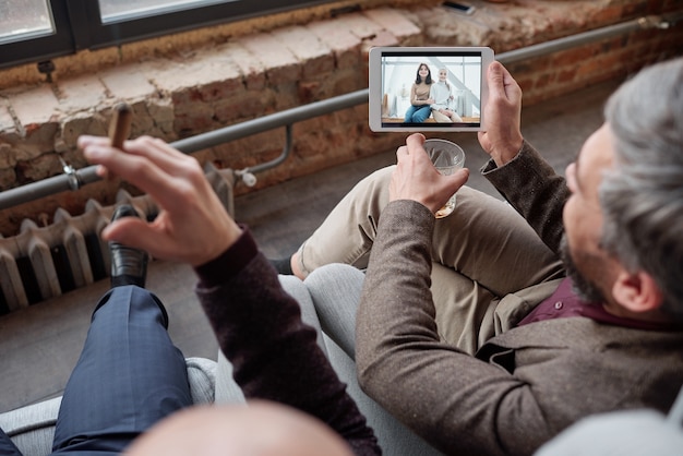 Vista por cima do ombro do empresário com o vidro compartilhando a foto da esposa e da filha no tablet para um amigo na festa