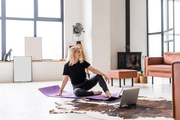 Vista por trás de uma mulher esportiva em roupas de fitness na moderna sala de estar usando o programa de treinamento de fitness on-line no laptop enquanto estava deitado no tapete de fitness.