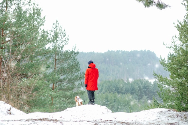 Vista posterior de un adolescente parado cerca de un acantilado entre pinos en el bosque del parque en un paseo nevado de invierno
