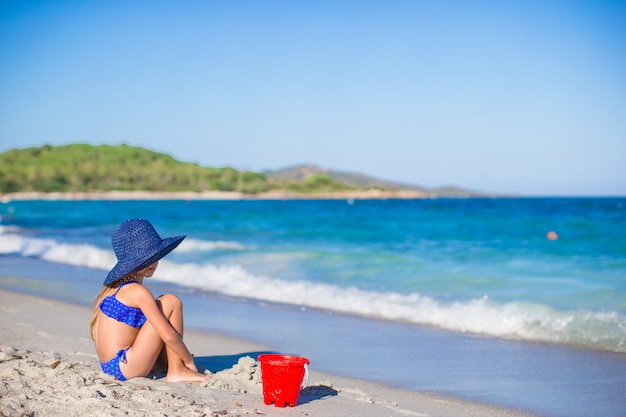 Vista posterior de la adorable niña en gran sombrero de paja azul en la playa blanca