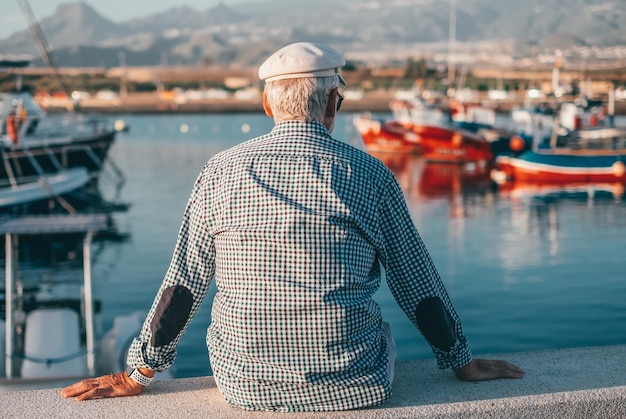 Vista posterior de un anciano relajado con camisa informal y sombrero sentado en el puerto marítimo al atardecer mirando los barcos Abuelo mayor disfrutando de viajes de tiempo libre o retiro en actividades de ocio