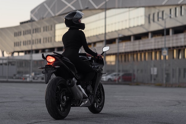 Vista posterior de la atractiva joven en traje negro ajustado y casco protector integral monta en motocicleta deportiva en el estacionamiento urbano al aire libre en la noche.