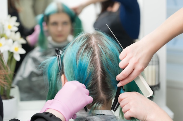 Vista posterior de la cabeza femenina con cabello color esmeralda y raíces capilares regeneradas. Mujer sentada en una silla junto al espejo, dos peluqueros peinando el cabello del cliente