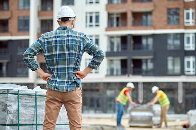 Vista posterior de un capataz en un casco de seguridad de pie con las manos en las caderas en el sitio de construcción