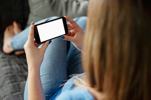 Vista posterior de cerca de una mano de mujer sosteniendo y usando un teléfono inteligente con pantalla vacía en blanco blanco sentado en una cama en casa