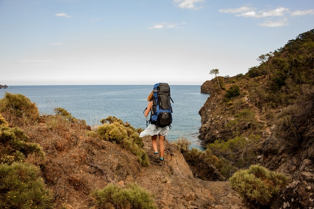 Vista posterior chica con una mochila de senderismo de pie sobre la roca en la costa