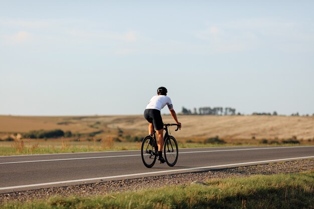 Vista posterior del ciclista profesional en blanco t-short y shorts negros montando bicicleta en carretera pavimentada