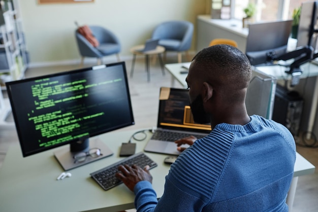 Vista posterior del desarrollador de software afroamericano escribiendo código en el lugar de trabajo en la oficina con múltiples d