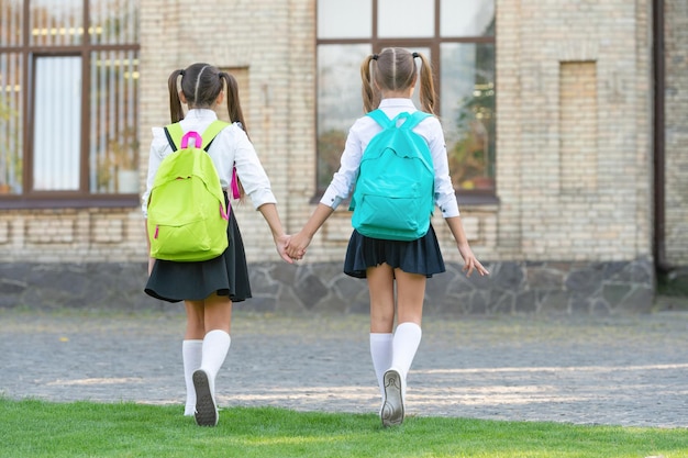 Vista posterior de dos colegialas con mochila escolar caminando juntos infancia al aire libre