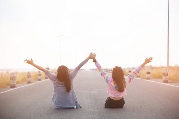 La vista posterior de dos mujeres jóvenes sentados en la carretera, mirando a lo lejos y las manos en alto.