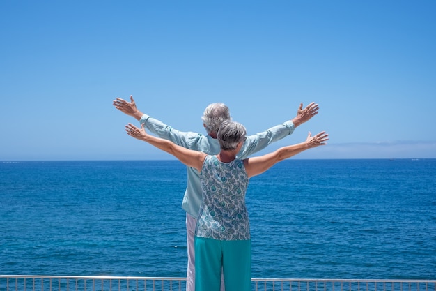 Vista posterior de dos personas mayores de pie en el mar con los brazos extendidos, mirando al horizonte disfrutando del verano y la libertad