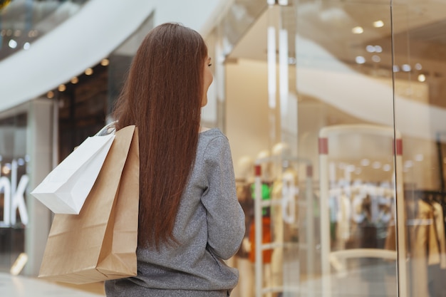 Vista posterior foto de una clienta en el centro comercial, sosteniendo bolsas de papel con la compra.