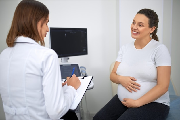 Foto vista posterior de la ginecóloga hablando y escribiendo en el portapapeles durante el chequeo médico de la joven embarazada