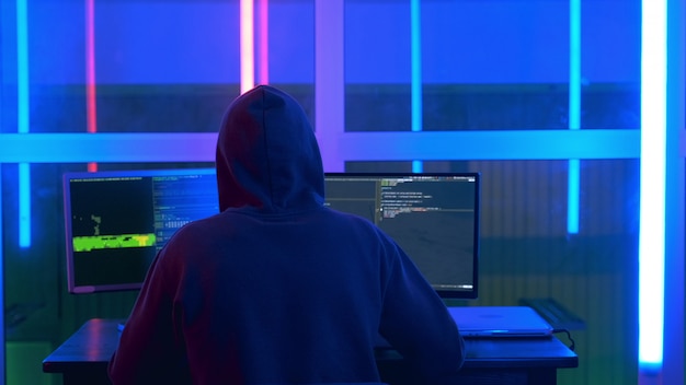 Vista posterior del hacker con una chaqueta negra y una capucha en la cabeza, escribiendo programas de hackers