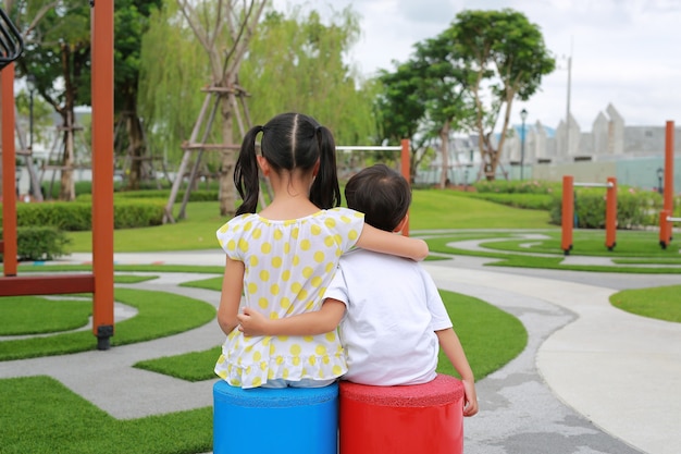 Vista posterior de la hermana mayor abraza al hermano pequeño por el cuello, los hombros sentados en el patio de recreo en el jardín con mirar al aire libre en el parque.