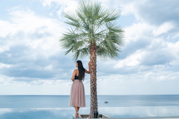 Vista posterior: una hermosa mujer de cabello largo con un vestido de verano se encuentra cerca de una palmera y una piscina infinita y admira la vista del mar