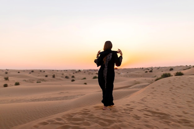 Vista posterior de Hermosa mujer misteriosa con vestido largo negro árabe tradicional se encuentra en el desierto al atardecer