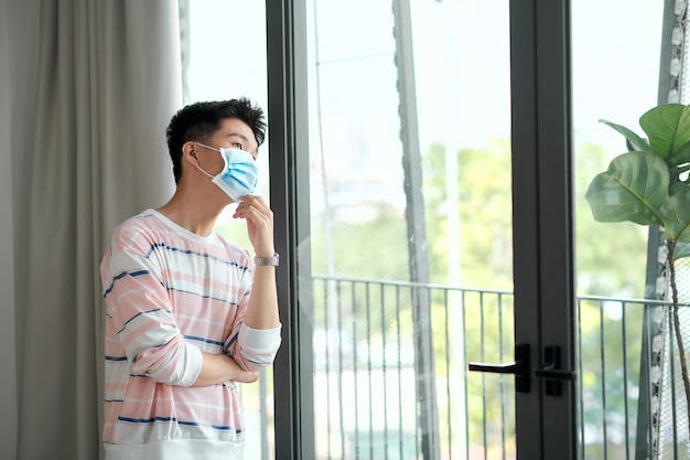 Vista posterior del joven asiático con mascarilla permanezca aislado en casa para auto cuarentena debido a la epidemia de COVID19 - él mira por la ventana