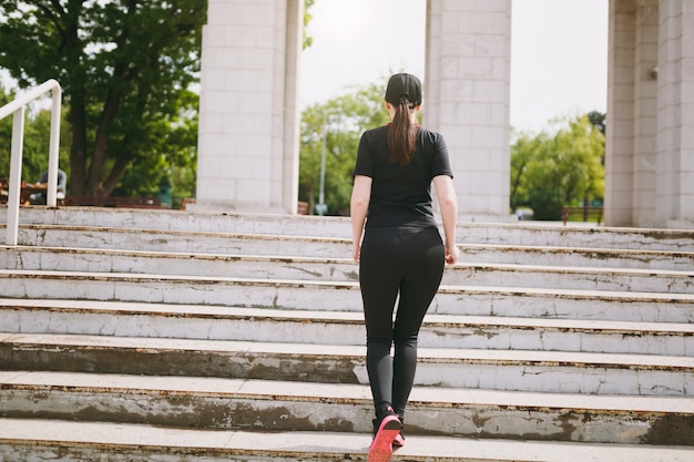 Vista posterior de la joven atlética fuerte morena en uniforme negro y gorra haciendo ejercicios deportivos, calentamiento antes de correr subiendo escaleras en el parque de la ciudad al aire libre