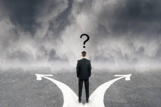 Vista posterior del joven empresario con signo de interrogación y dos flechas en la carretera de hormigón Fondo de cielo nublado Dirección diferente y concepto de confusión