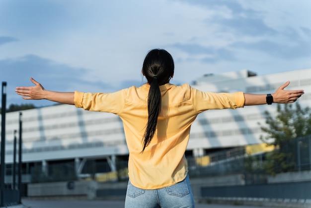 Vista posterior de la joven extendiendo las manos con alegría e inspiración frente al sol. Mujer respirando aire fresco y estirando los brazos en la calle