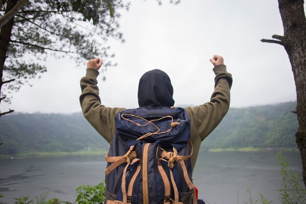 Vista posterior de la joven mujer asiática musulmana con las manos en alto de pie sobre un acantilado sobre el lago. Concepto de viaje, conceptos de ganador, libertad