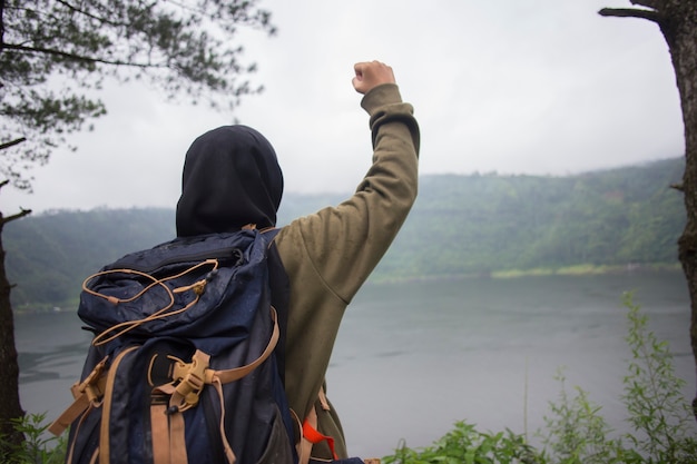 Vista posterior de la joven mujer asiática musulmana con las manos en alto de pie sobre un acantilado sobre el lago. Concepto de viaje, conceptos de ganador, libertad